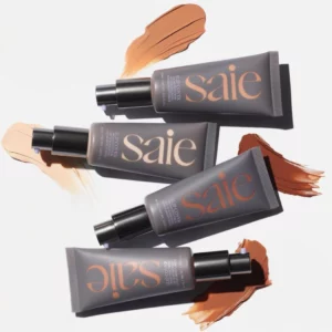 Cztery tubki kremu koloryzującego Saie Slip Tint SPF 35 ułożone na białym tle, każda z rozsmarowaną próbką produktu w różnych odcieniach – od bardzo jasnego po głęboki, ciepły brąz. Tubki mają matowe, grafitowe opakowania z dużym napisem „saie” i pompką dozującą.