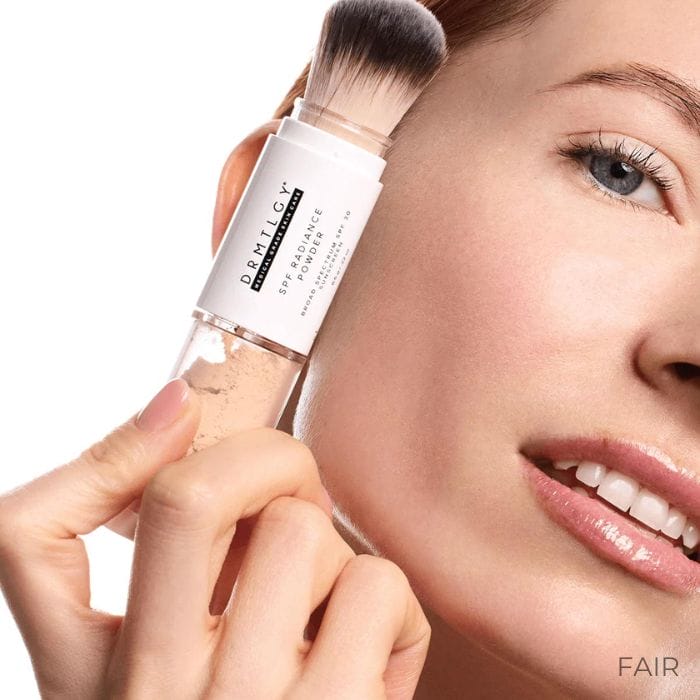 Drmtlgy-SPF-Radiance-Powder-Fair3 Na obrazku znajduje się zbliżenie twarzy kobiety, która nakłada puder DRMTLGY Radiance z SPF 50 na policzek za pomocą pędzelka z wbudowanym aplikatorem. Kosmetyk ma kolor jasny, oznaczony jako "Fair". Kobieta ma jasną karnację i delikatny uśmiech.