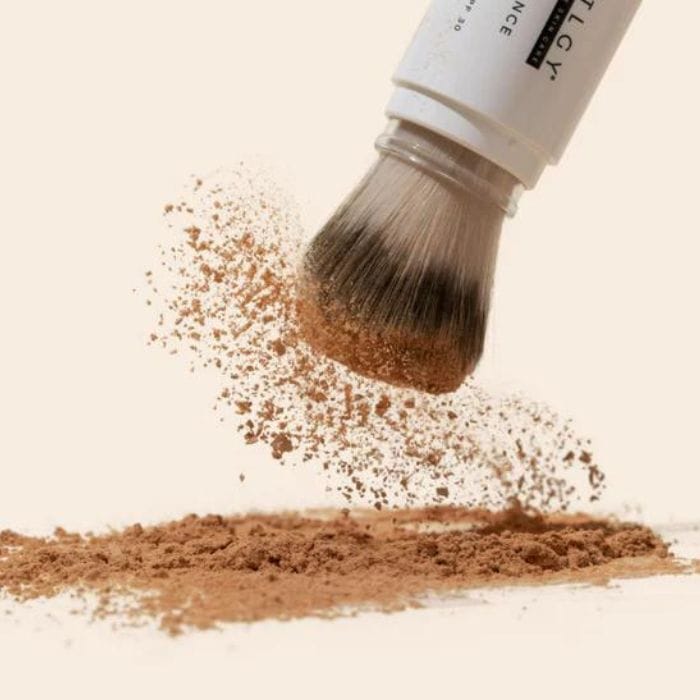 Drmtlgy-SPF-Radiance-Powder5 Na tym zdjęciu przedstawiony jest pędzel marki Drmtlgy w trakcie aplikacji pudru ochronnego SPF Radiance Powder. Pędzel widoczny jest z bliska, a puder sypie się z jego włosia, tworząc chmurę pudrową. Produkt ten łączy właściwości ochrony przeciwsłonecznej SPF 30 z funkcją kosmetyku do makijażu, zapewniając wyrównany koloryt skóry i ochronę przed szkodliwym działaniem promieni UV. Puder jest w odcieniu beżu, a pędzel posiada biały uchwyt z napisami "DRMTLGY" oraz opisem produktu.