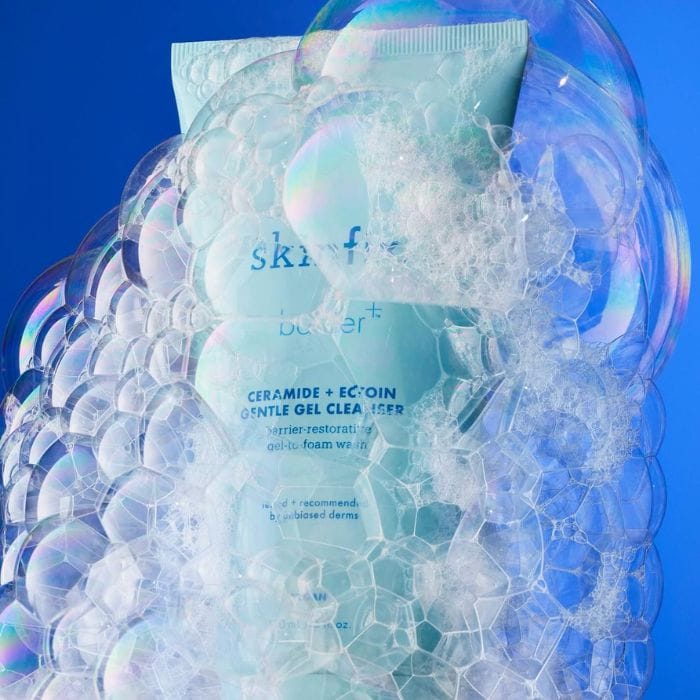 Skinfix-Barrier-Gentle-Gel-Cleanser6 Na obrazku znajduje się tubka produktu Skinfix Barrier+ Ceramide + Ectoin Gentle Gel Cleanser, zanurzona w gęstej pianie mydlanej. Tubka ma jasno niebieski kolor i jest częściowo ukryta za bąbelkami, co podkreśla właściwości oczyszczające i pieniące produktu. Napis na tubce jest widoczny mimo bąbelków i zawiera informacje o produkcie: "skinfix" "barrier+" "CERAMIDE + ECTOIN GENTLE GEL CLEANSER" "barrier-restorative gel-to-foam wash" "tested + recommended by unbiased derms" "VEGAN" "150 mL / 5 fl. oz." Obrazek jest utrzymany w świeżej, czystej estetyce, z dominującym niebieskim tłem, co wzmacnia wrażenie czystości i skuteczności myjącej produktu.