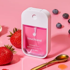 Na zdjęciu znajduje się mgiełka do dezynfekcji rąk marki Touchland w w przezroczystym, opakowaniu z białym dozownikiem i napisem "Berry Bliss". Na tle widoczne są truskawki, jagody oraz łyżeczka z syropem, co sugeruje owocowy aromat produktu.