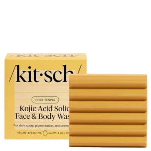 Zbliżenie na opakowanie mydła Kitsch "Kojic Acid Solid Face & Body Wash" oraz samo mydło w kolorze musztardowym. Mydło ma charakterystyczną, żłobioną strukturę, a opakowanie informuje o jego właściwościach rozjaśniających skórę.