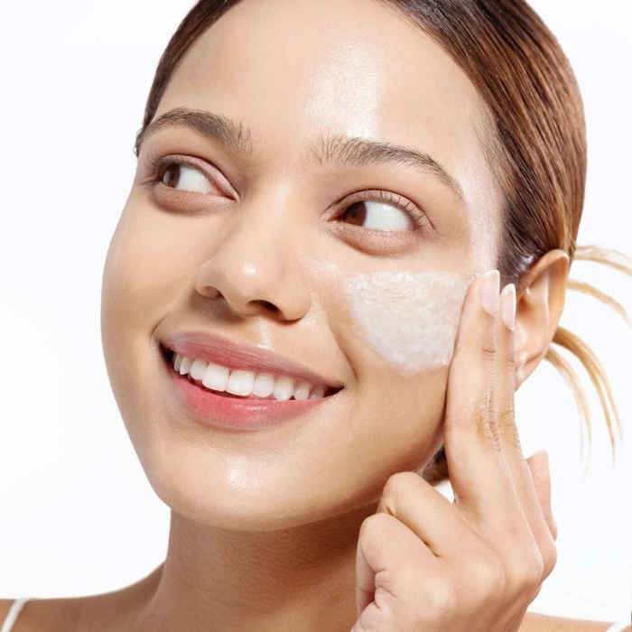 Arocell Super Collagen Melting Cleansing Balm Zdjęcie przedstawia uśmiechniętą kobietę, która nakłada na swoją twarz kosmetyk przypominający balsam. Produkt jest rozprowadzany na policzku, co sugeruje etap pielęgnacji skóry, jakim jest oczyszczanie lub nawilżanie. Skóra kobiety wygląda na zdrową, gładką i promienną, co podkreśla efekt stosowania produktu. Tło jest jasne, co nadaje zdjęciu świeżości i czystości.