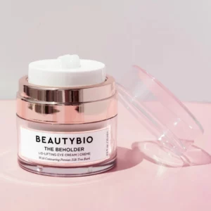 Elegancki słoiczek kremu BeautyBio The Beholder na różowym tle. Opakowanie ma luksusowy design z przezroczystymi i metalicznymi akcentami, podkreślając wysoką jakość produktu.