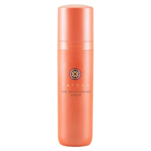 Na zdjęciu znajduje się zamknięta buteleczka serum rozjaśniającego marki Tatcha w eleganckim, pomarańczowym opakowaniu z minimalistycznym designem.