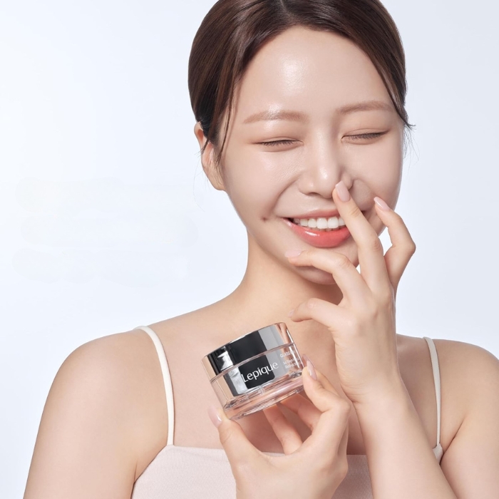 Lepique Glass Skin Retinol Night Cream (3) Na zdjęciu znajduje się kobieta o promiennej cerze, trzymająca słoiczek kremu Lepique Glass Skin 14 Days Miracle Retinol Night Cream. Kobieta ma uśmiechnięty wyraz twarzy, co sugeruje zadowolenie z produktu. Skóra wygląda na zdrową, nawilżoną i pełną blasku. Słoiczek kremu, utrzymany w eleganckim i minimalistycznym stylu, z przezroczystym opakowaniem i srebrnym wieczkiem, podkreśla luksusowy charakter produktu. Jasne tło i delikatny styl zdjęcia eksponują piękno skóry oraz wyjątkowość kremu dedykowanego nocnej pielęgnacji.