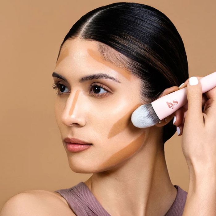 Lys Beauty Sculpting Brush (2) Kobieta z ciemnymi, gładko zaczesanymi włosami nakłada makijaż przy użyciu pędzla do konturowania. Jej twarz ma nałożone jaśniejsze i ciemniejsze odcienie kosmetyków w celu podkreślenia rysów. Pędzel, który trzyma dłoń, ma różowy trzonek i biało-czarne włosie.