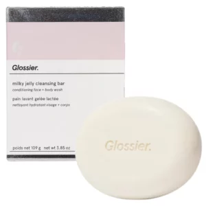 Produkt Glossier Milky Jelly Cleansing Bar: owalne, białe mydło z wytłoczoną nazwą marki „Glossier” na powierzchni. W tle widoczne jest pastelowe opakowanie w odcieniach różu, fioletu i bieli z napisem „silky jelly cleansing bar”.