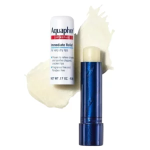 Zdjęcie przedstawia Aquaphor Lip Repair, ochronny balsam do ust w sztyfcie. Widoczny jest zarówno zamknięty produkt w białym opakowaniu z etykietą, jak i otwarty sztyft w metalicznym granatowym opakowaniu, odsłaniający beżowy balsam. W tle znajduje się kremowa warstwa produktu, podkreślająca jego bogatą, nawilżającą konsystencję. Na etykiecie widnieją informacje o właściwościach balsamu – natychmiastowa ulga dla bardzo suchych ust, udowodniona skuteczność w łagodzeniu suchości, spierzchniętych i popękanych ust, brak substancji zapachowych i parabenów oraz waga produktu: 4,8 g.