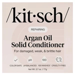 Zdjęcie opakowania Kitsch Argan Oil Solid Conditioner – odżywki w kostce z olejem arganowym, przeznaczonej do włosów zniszczonych, osłabionych i łamliwych. Minimalistyczny design w beżowych odcieniach zawiera czarne napisy z nazwą marki i produktu oraz informację, że jest to regenerująca formuła ("Repairing"). Na dole opakowania znajdują się ikony i oznaczenia wskazujące, że produkt jest bezpieczny dla włosów farbowanych (Color Safe), ma zrównoważone pH, wystarcza na około 100 użyć i jest cruelty-free. Waga produktu to 77 g.