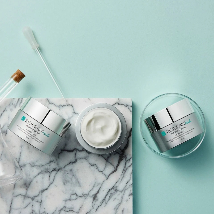 rejuran healer cream (5) Stylowe ujęcie słoiczków REJURAN Nutritive Cream na marmurowej podstawce, otoczonej pipetą i szklanym naczyniem, na pastelowym tle.