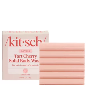 Kostka i opakowanie Kitsch Tart Cherry Solid Body Wash ułożone na różowym tle obok rozciętych i całych wiśni oraz rozlanej cieczy w odcieniu różu.