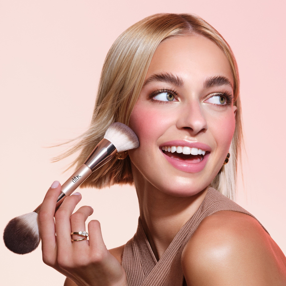 Dibs-Duo-Brush-Face (2) Kobieta z krótkimi blond włosami nakłada róż na policzki pędzlem Dibs Duo Brush Face, uśmiechając się szeroko.
