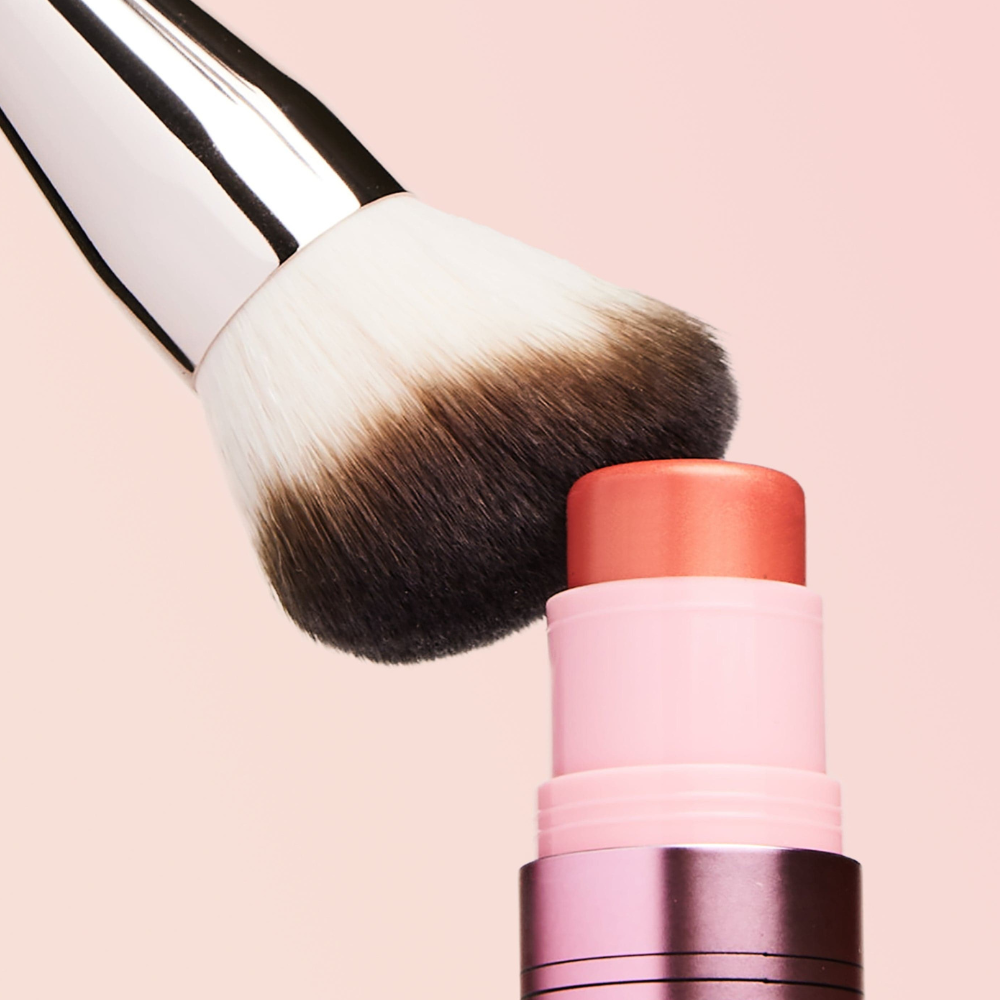 Dibs-Duo-Brush-Face Zbliżenie pędzla Dibs Duo Brush Face dotykającego różu w sztyfcie w odcieniu koralowym.