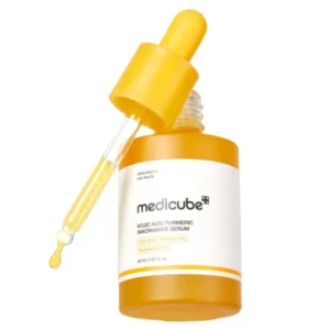 Pionowo ustawiona i otwarta butelka serum Medicube Kojic Acid Turmeric Niacinamide Serum w intensywnie żółtym kolorze, z której wypływa kropla produktu. Obok widoczna jest pipeta pokryta złocistym, gęstym serum.