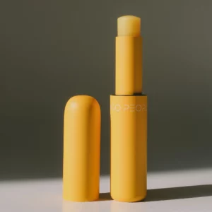 Żółty balsam do ust Mango People w minimalistycznym opakowaniu typu stick, wysunięta formuła na tle ciepłego światła.