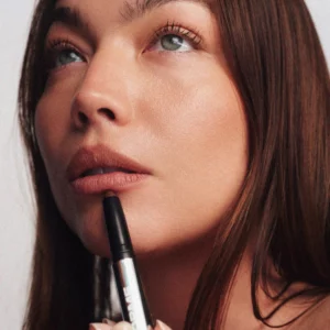Modelka z naturalnym makijażem ust w odcieniu Teddy, trzymająca pomadkę Reforme Power Lip Sculpting Wand przy wargach, prezentująca miękkie, matowe wykończenie.