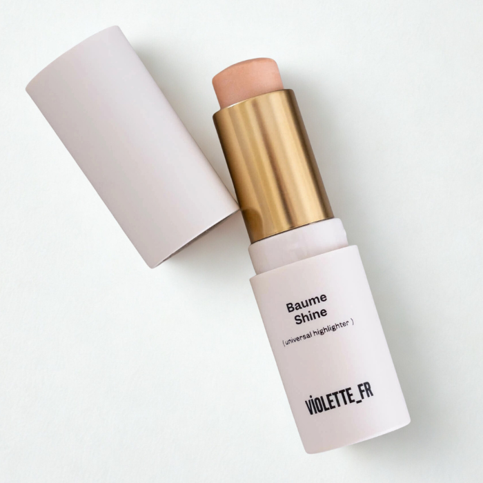 4 Zbliżenie na rozświetlacz w sztyfcie Baume Shine (universal highlighter) marki Violette_FR. Produkt w jasnym, minimalistycznym opakowaniu z białą nakrętką i złotym wnętrzem, wysunięty w odcieniu delikatnego beżu z subtelnym połyskiem. Ujęcie na jasnym tle podkreśla elegancki, francuski charakter kosmetyku.