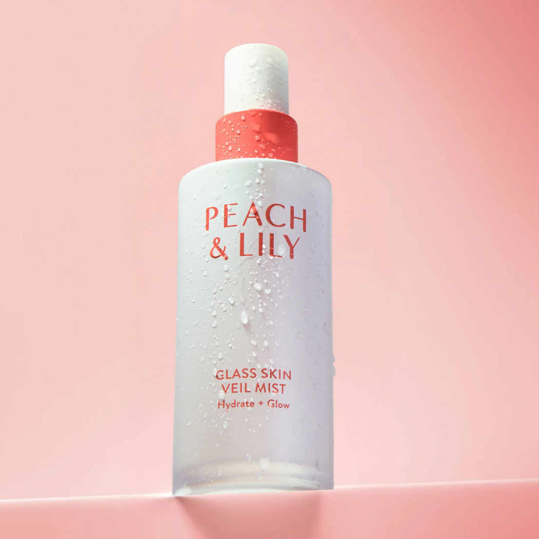 Peach Lily Glass Skin Veil Mist (2) Buteleczka Peach & Lily Glass Skin Veil Mist pokryta kroplami wody na tle pastelowego różu, symbolizująca świeżość i nawilżenie skóry.
