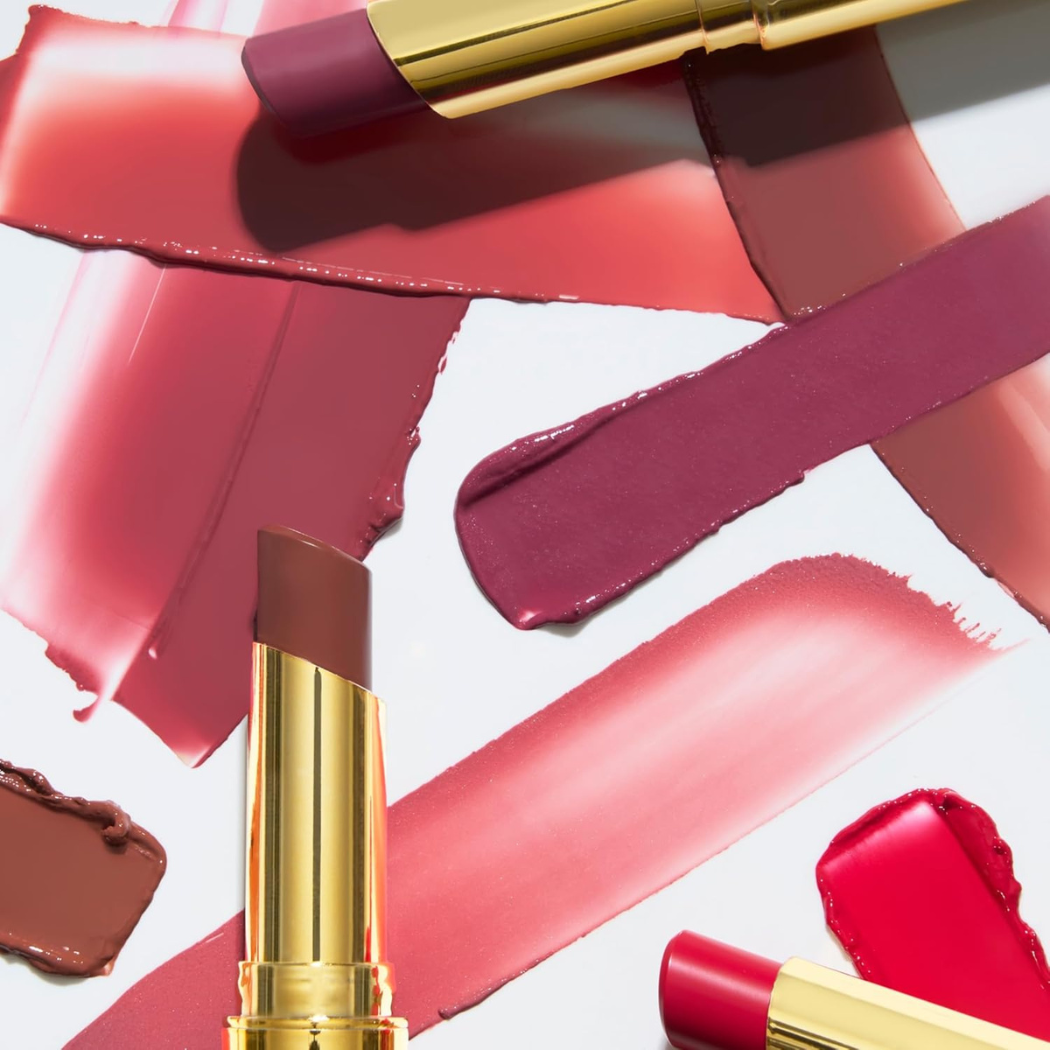 Laura Geller Jelly Balm Hydrating Lip Color Kilka smug balsamu do ust w różnych odcieniach różu, beżu i brązu, rozprowadzonych na białym tle. Wśród nich widoczne złote opakowania balsamu z wysuniętym produktem.