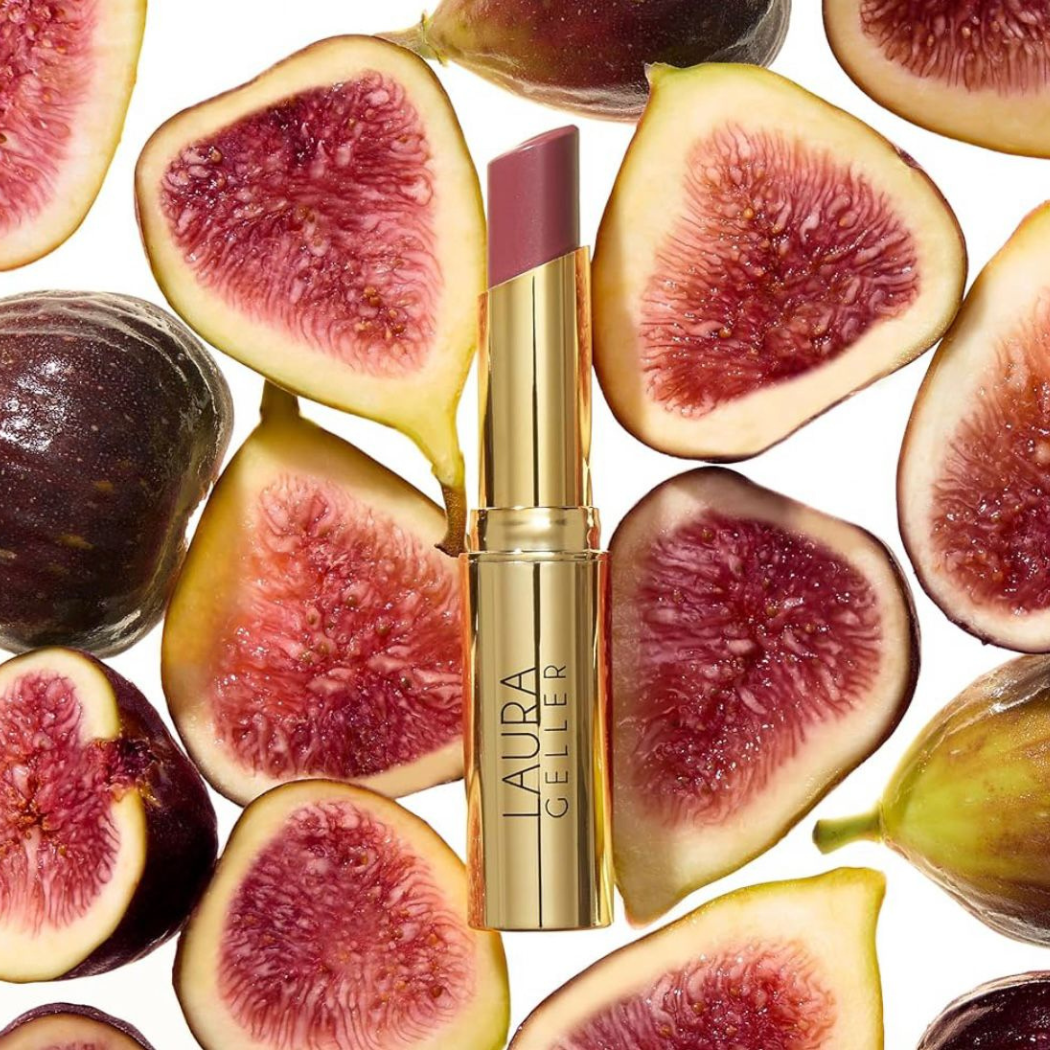Laura Geller Jelly Balm Hydrating Lip Color Złote opakowanie balsamu do ust ustawione pionowo, otoczone połówkami świeżych fig o czerwono-różowym wnętrzu. Tło jasne, kompozycja dekoracyjna.