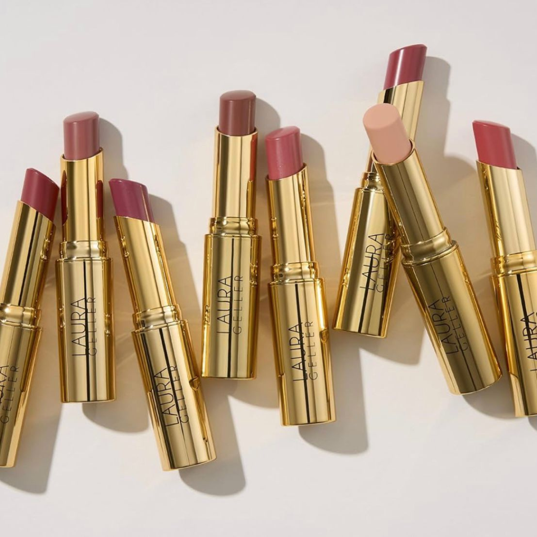 Laura Geller Jelly Balm Hydrating Lip Color Kilka otwartych balsamów do ust w złotych opakowaniach, ułożonych obok siebie na jasnym tle. Widoczne różne odcienie produktu, od jasnych po ciemniejsze.