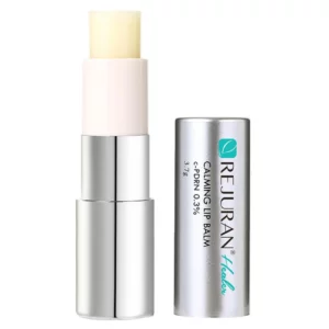 Balsam do ust Rejuran Calming Lip Balm w sztyfcie z widoczną zawartością w kolorze kremowym, w eleganckim opakowaniu w kolorze srebrnym i białym. Obok stoi zdejmowana nakrętka z logo i opisem produktu.