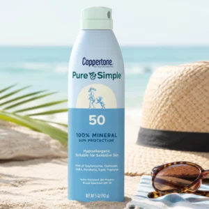 Minimalistyczny packshot mineralnego sprayu przeciwsłonecznego Coppertone Pure & Simple SPF 50 na plaży. Obok produktu znajdują się kapelusz przeciwsłoneczny, okulary i liść palmowy, podkreślające wakacyjny charakter produktu.