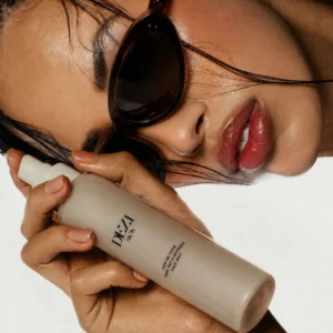 Dezi Skin Dew Me Over Face Mist – modelka w okularach przeciwsłonecznych prezentuje odświeżającą mgiełkę do twarzy o lekkiej, nawilżającej formule.