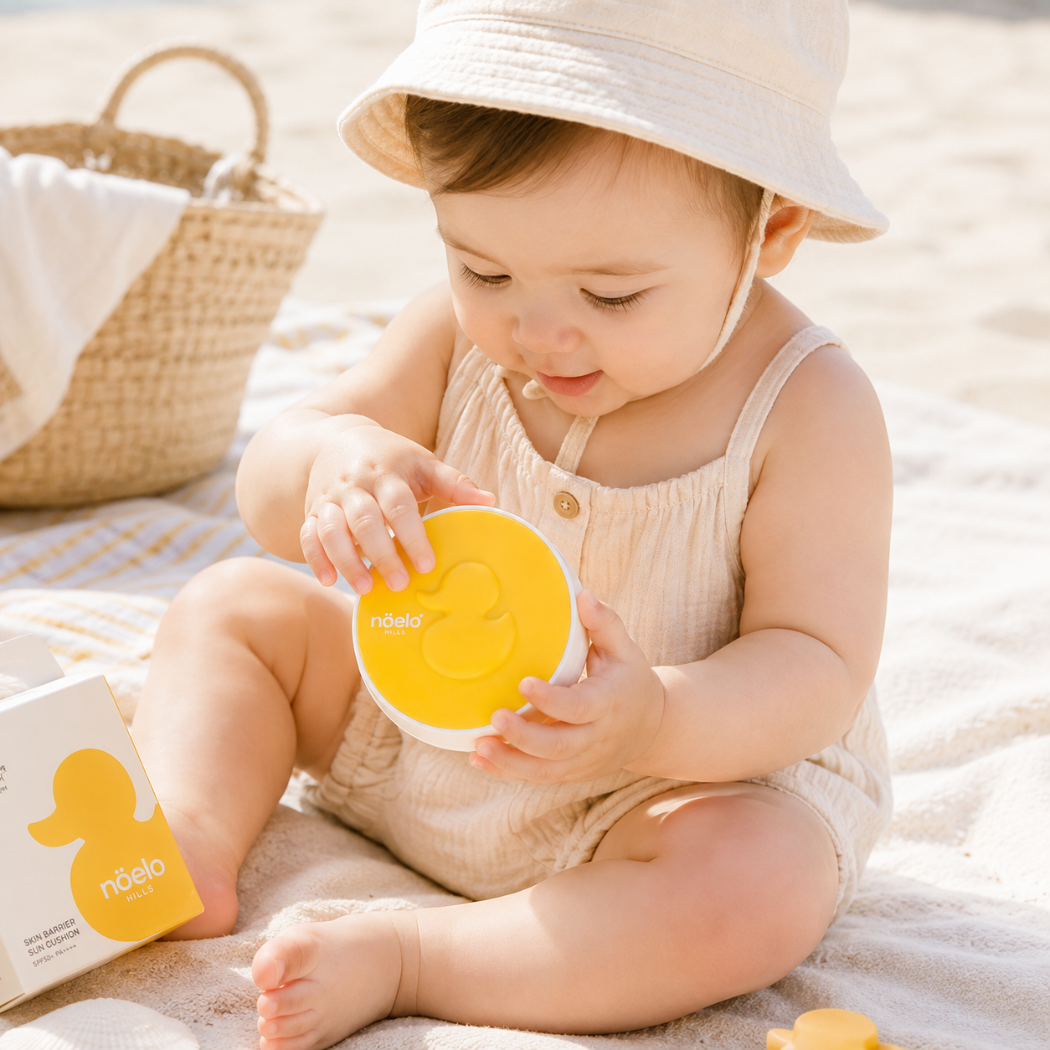 Noelo Hills Skin Barrier Sun Cushion SPF 50 Na zdjęciu znajduje się małe dziecko siedzące na jasnym kocu na plaży, trzymające w dłoniach żółty cushion przeciwsłoneczny Noelo Hills Skin Barrier Sun Cushion SPF50+ PA++++. Obok widoczne jest opakowanie produktu oraz pleciony kosz, a całość utrzymana jest w ciepłej, wakacyjnej estetyce.