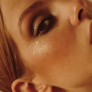 Na zdjęciu widoczne są złote aplikacje Fazit Golden Hour Eye Wings zaaplikowane pod okiem. Delikatne linie, kropki i gwiazdka rozświetlają makijaż, nadając mu festiwalowy, biżuteryjny efekt.