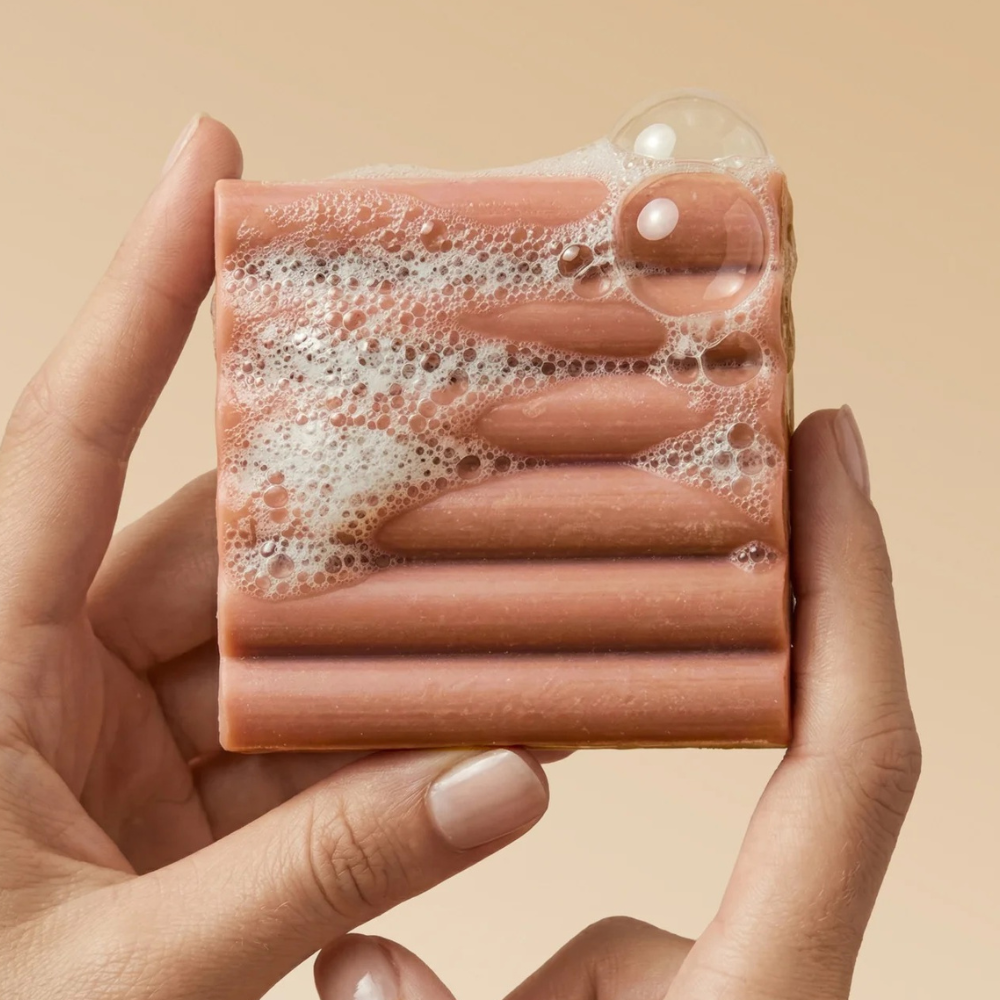 Kitsch Amber Shores Body Wash Bar Kitsch Amber Shores Body Wash Bar – kostka myjąca do ciała trzymana w dłoniach, pokryta kremową pianą i bąbelkami.