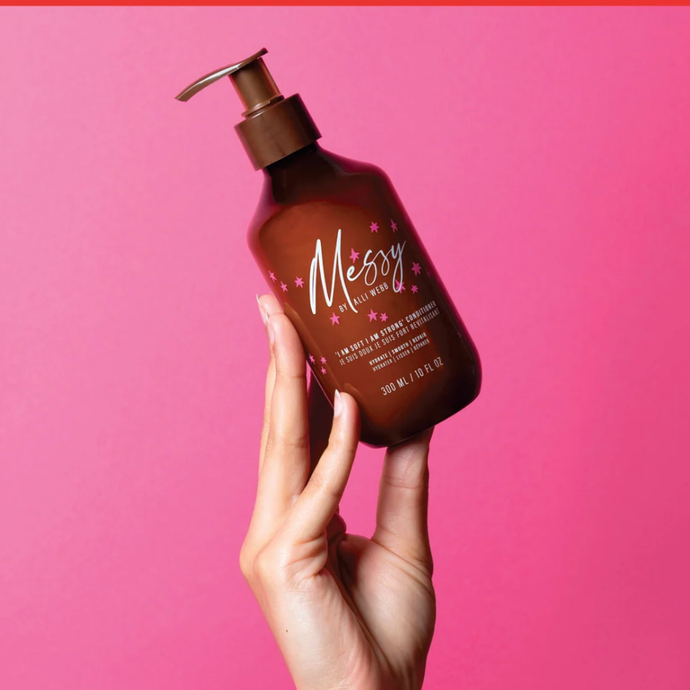 Messy by Alli Webb Hydrating & Cleansing Shampoo Dłoń trzyma brązową butelkę odżywki Messy by Alli Webb na intensywnie różowym tle.