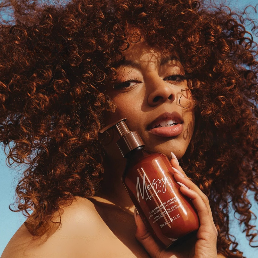 Messy by Alli Webb Hydrating & Cleansing Shampoo Modelka z burzą naturalnych loków trzyma w dłoni brązową butelkę Messy by Alli Webb na tle błękitnego nieba.