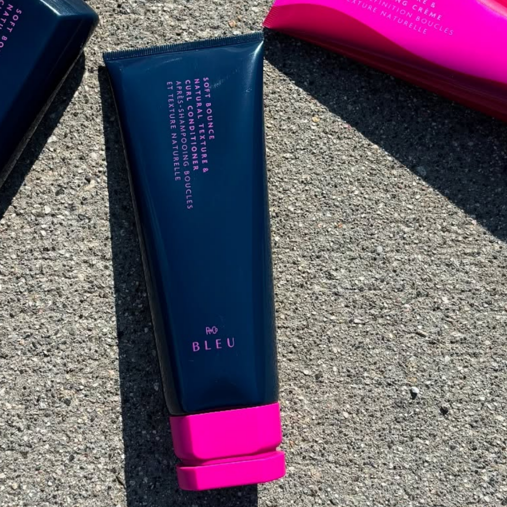 R+Co BLEU Soft Bounce Natural Texture & Curl Conditioner Odżywka R+Co BLEU Soft Bounce Natural Texture & Curl Conditioner ułożona na betonowej powierzchni w słońcu, obok pozostałych kosmetyków z kolekcji.