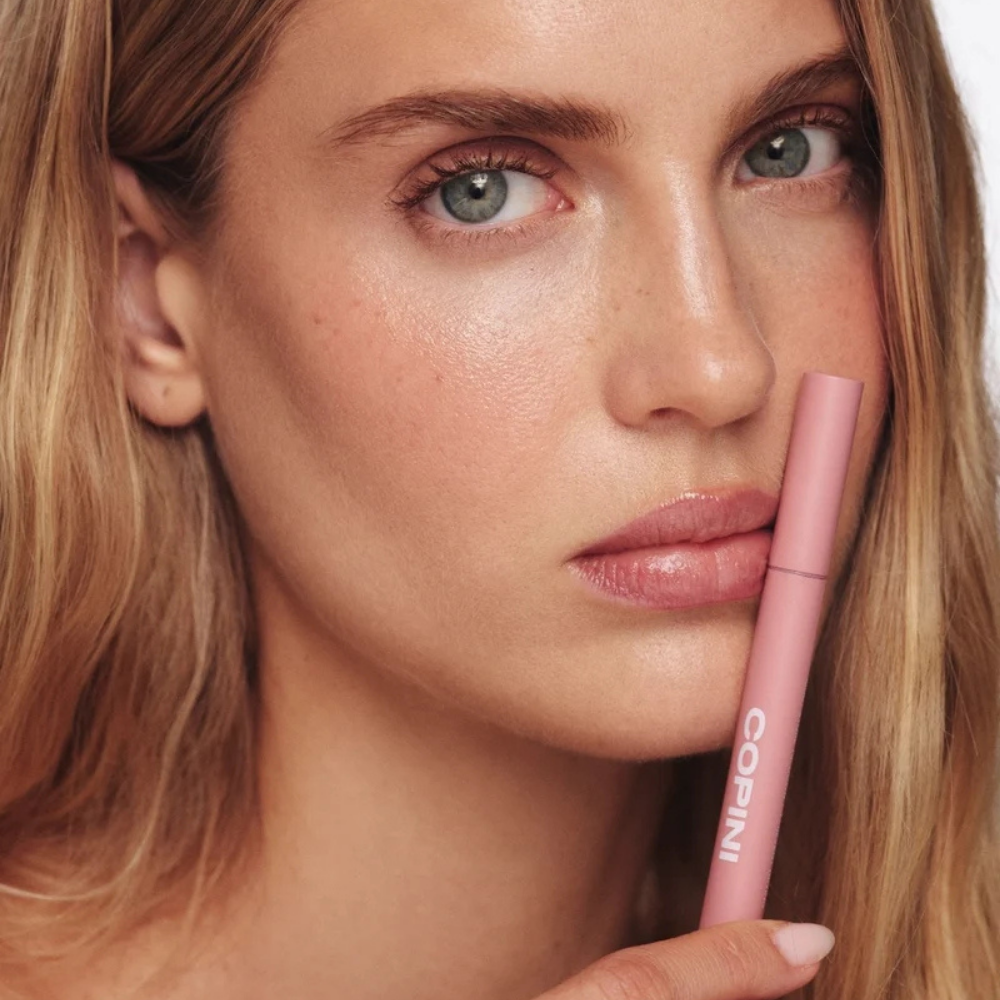 Copini Love Bite Soft Focus Lip Stain (3) Copini Love Bite Soft Focus Lip Stain - efekt Pillow - modelka prezentująca subtelny różowy odcień Pillow na ustach oraz różowe opakowanie produktu.