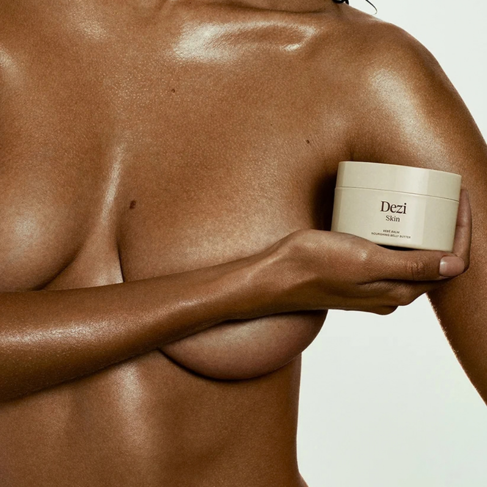 Dezi Skin Bebe Balm - odżywcze masło do ciała i brzucha (2) Dezi Skin Bebé Balm - odżywcze masło do ciała trzymane w dłoni na tle nawilżonej, promiennej skóry ciała.