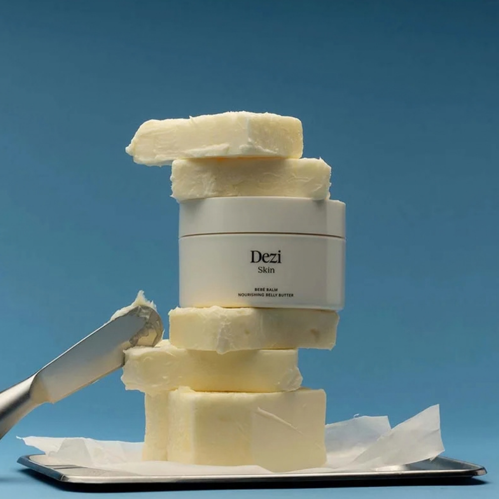Dezi Skin Bebe Balm - odżywcze masło do ciała i brzucha Dezi Skin Bebé Balm - bogata maślana formuła: opakowanie odżywczego masła do ciała otoczone kawałkami masła na niebieskim tle, podkreślające jego kremową, odżywczą konsystencję.