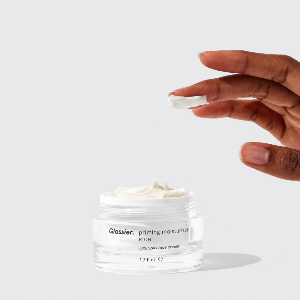 Glossier Priming Moisturizer Rich - otulający sekret gładkiej cery (2) Glossier Priming Moisturizer Rich - otwarty słoiczek z bogatym kremem do twarzy oraz dłoń prezentująca jego gęstą, kremową konsystencję.