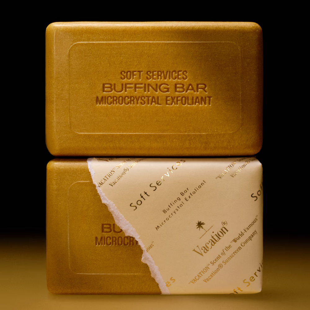 Soft Services X Vacation Buffing Bar Exfoliant - kostka peelingująca pachnąca jak najlepsze wakacje (2) Soft Services x Vacation Buffing Bar - mikrokrystaliczna kostka peelingująca - dwie złociste kostki peelingujące do ciała Soft Services x Vacation, w tym jedna częściowo owinięta papierem z logo Vacation, na czarnym tle.