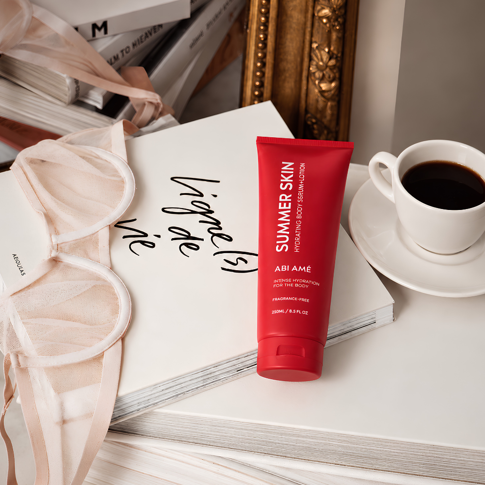 Abi Ame Summer Skin (2) Czerwone serum-lotion do ciała Summer Skin marki ABI AMÉ, bezzapachowe, o pojemności 250 ml, ustawione na otwartej książce obok jasnoróżowej bielizny i filiżanki czarnej kawy. Elegancka aranżacja w ciepłych, neutralnych odcieniach.