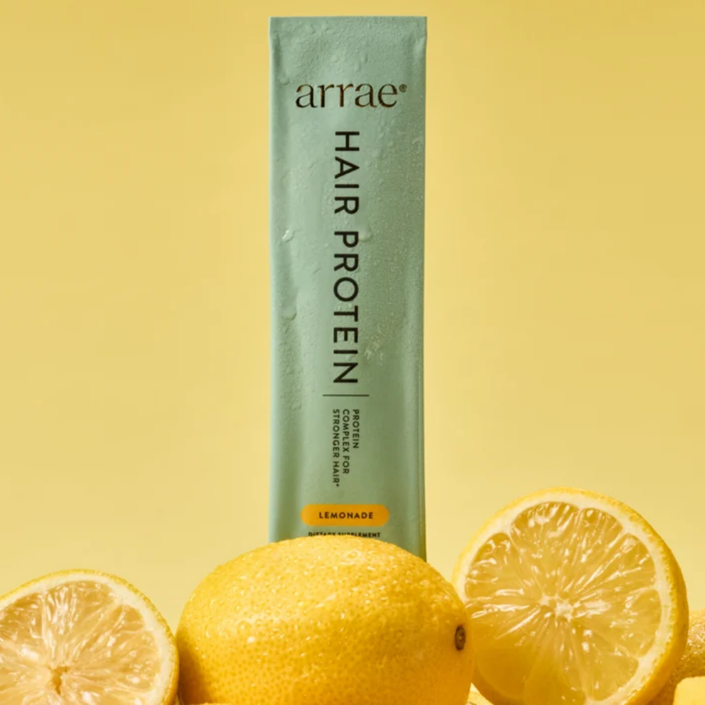 Arrae Hair Protein Hair Growth Stick Packs Lemonade Arrae Hair Protein Hair Growth Stick Packs Lemonade - saszetka suplementu diety wspierającego kondycję i zdrowy wygląd włosów, w miętowozielonym opakowaniu, otoczona świeżymi cytrynami na pastelowym żółtym tle.