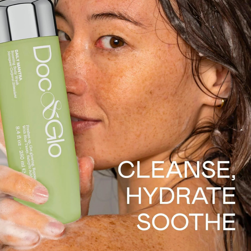 Doc & Glo Daily Mantra Boosting Body Wash (4) Kobieta z piegami i mokrymi, ciemnymi włosami trzyma przy twarzy jasnozieloną butelkę Doc & Glo Daily Mantra Boosting Body Wash o pojemności 250 ml. Dłonie pokryte delikatną pianą, a na grafice biały napis „Cleanse, Hydrate, Soothe” podkreśla oczyszczające, nawilżające i kojące właściwości żelu do mycia ciała.