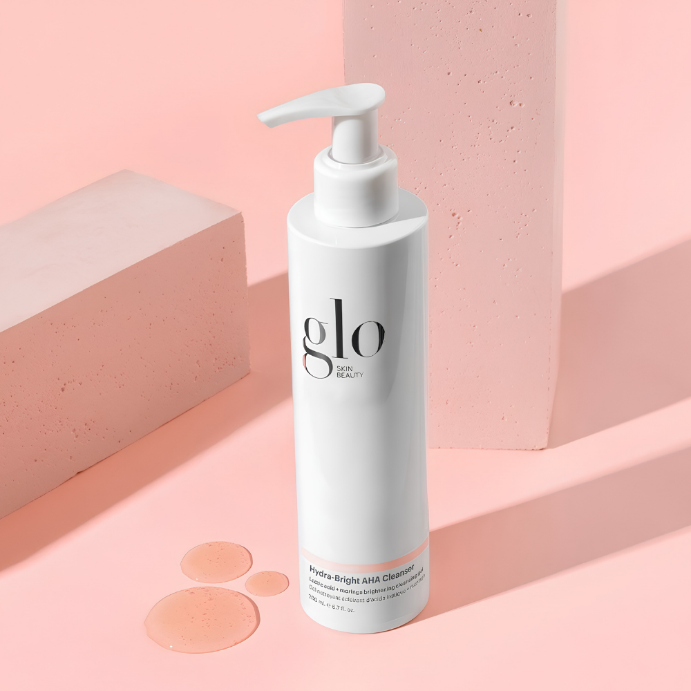 Glo Skin Beauty Hydra Bright AHA Cleanser (2) Glo Skin Beauty Hydra-Bright AHA Cleanser 200 ml - rozświetlający żel do mycia twarzy z kwasem mlekowym AHA i moringą. Biała butelka z pompką na pastelowym, różowym tle, otoczona geometrycznymi bryłami i kroplami żelu. Kosmetyk delikatnie oczyszcza, wygładza skórę i pomaga przywrócić cerze naturalny blask.