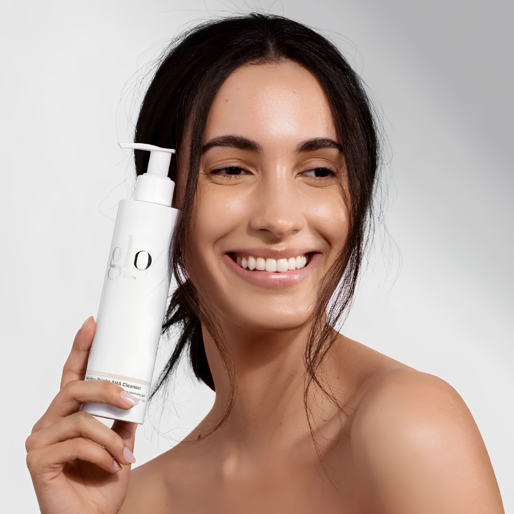 Glo Skin Beauty Hydra Bright AHA Cleanser (3) Glo Skin Beauty Hydra-Bright AHA Cleanser 200 ml - rozświetlający żel do mycia twarzy z kwasem mlekowym AHA i moringą. Uśmiechnięta modelka o ciemnych włosach i promiennej cerze trzyma białą butelkę z pompką przy twarzy. Kosmetyk pomaga delikatnie złuszczać martwy naskórek, wygładzać skórę i przywracać jej naturalny blask.