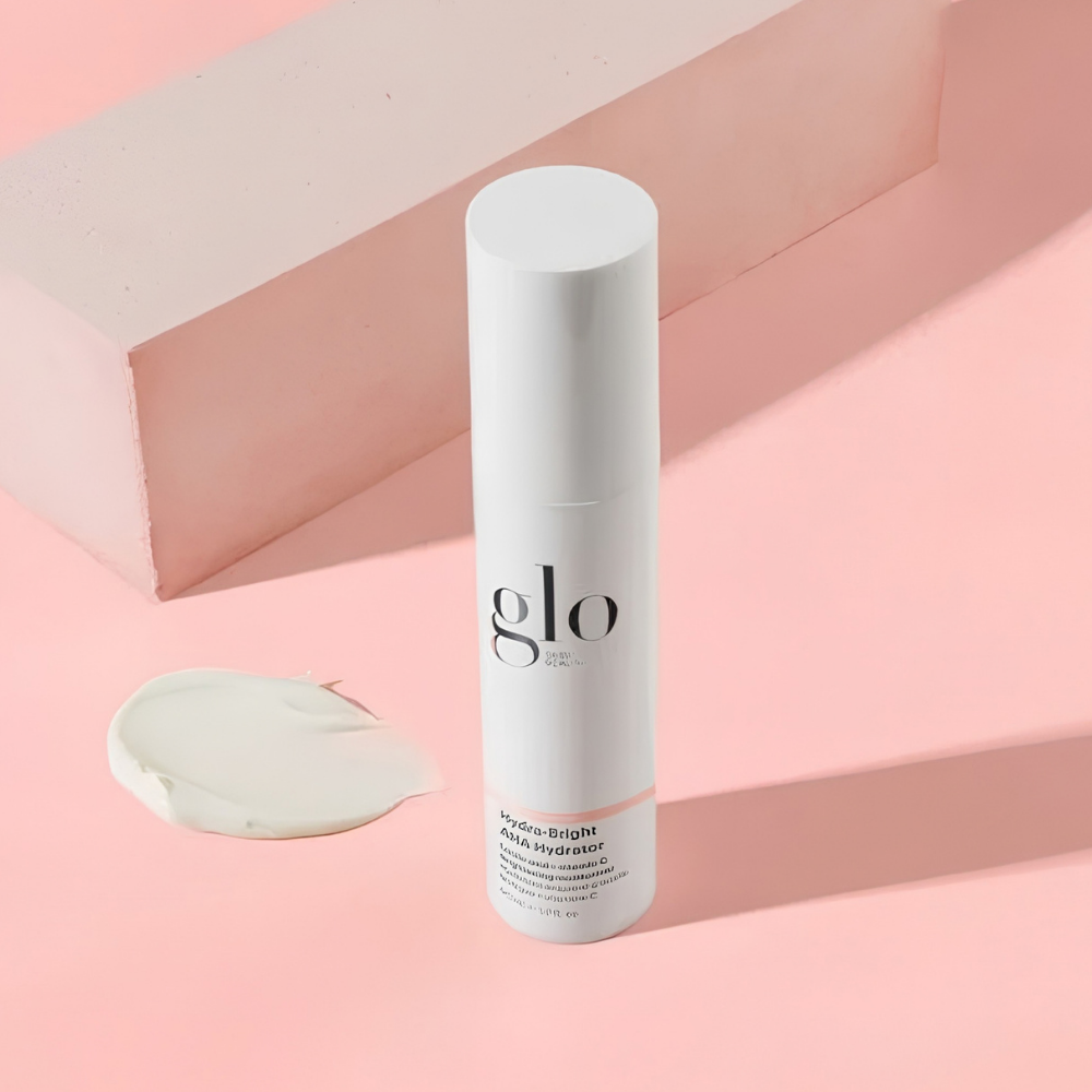 Glo Skin Beauty Hydra Bright AHA Hydrator (2) Glo Skin Beauty Hydra-Bright AHA Hydrator - nawilżający krem rozświetlający z kwasami AHA i witaminą C, 50 ml. Elegancka biała butelka na pasteloworóżowym tle, obok widoczna kremowa konsystencja kosmetyku oraz geometryczne elementy dekoracyjne.