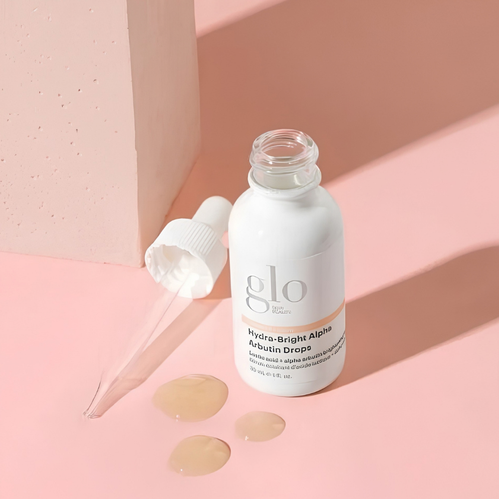 Glo Skin Beauty Hydra Bright Alpha Arbutin Drops (3) Glo Skin Beauty Hydra-Bright Alpha Arbutin Drops - serum rozświetlające z arbutyną i kwasami AHA na przebarwienia, 30 ml. Otwarta biała buteleczka z logo marki, szklana pipeta i trzy krople serum o lekkiej, żelowej konsystencji na pasteloworóżowym tle z geometrycznymi dekoracjami.