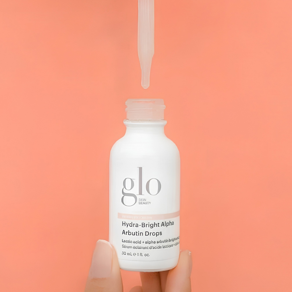 Glo Skin Beauty Hydra Bright Alpha Arbutin Drops (4) Glo Skin Beauty Hydra-Bright Alpha Arbutin Drops - serum rozświetlające z arbutyną i kwasami AHA na przebarwienia, 30 ml. Biała buteleczka z logo marki trzymana w dłoni, nad nią szklana pipeta z kroplą serum, na intensywnie koralowym tle.