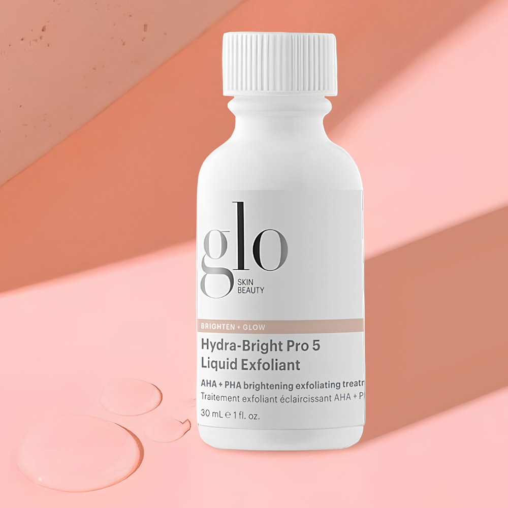 Glo Skin Beauty Hydra-Bright Pro 5 Liquid Exfoliant Glo Skin Beauty Hydra-Bright Pro 5 Liquid Exfoliant 30 ml - rozświetlający peeling do twarzy z kwasami AHA i PHA, wspomagający wygładzenie skóry i wyrównanie kolorytu. Biała butelka na eleganckim, brzoskwiniowo-różowym tle z kroplami transparentnego płynu.