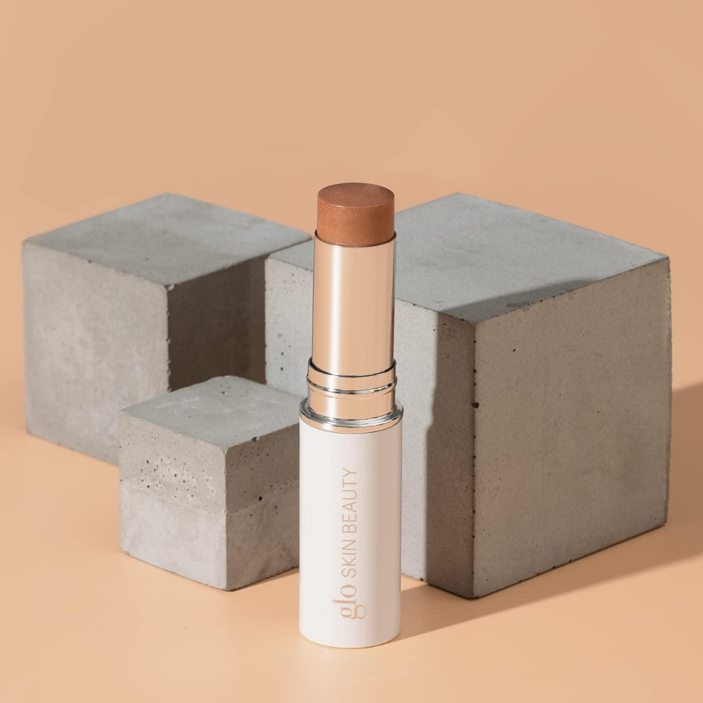 Glo Skin Beauty Skin Glow Stick Highlighter (4) Glo Skin Beauty Skin Glow Stick Highlighter - kremowy rozświetlacz w sztyfcie w ciepłym, brązowo-złotym odcieniu, otwarte biało-srebrne opakowanie na brzoskwiniowym tle z betonowymi podestami, kosmetyk do naturalnego rozświetlania twarzy.